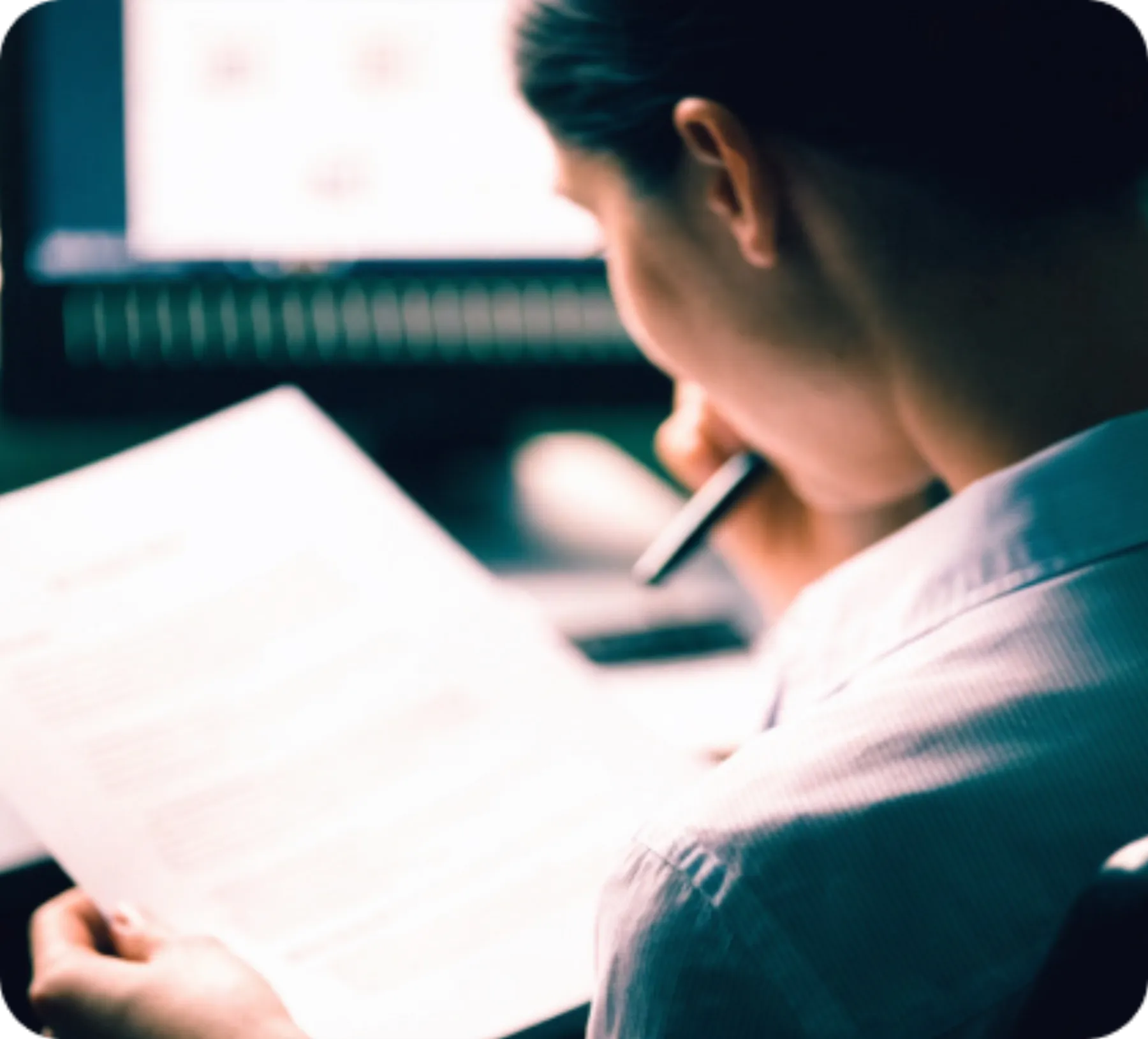 Someone at a desk reading a printed page, a screen out of focus behind them