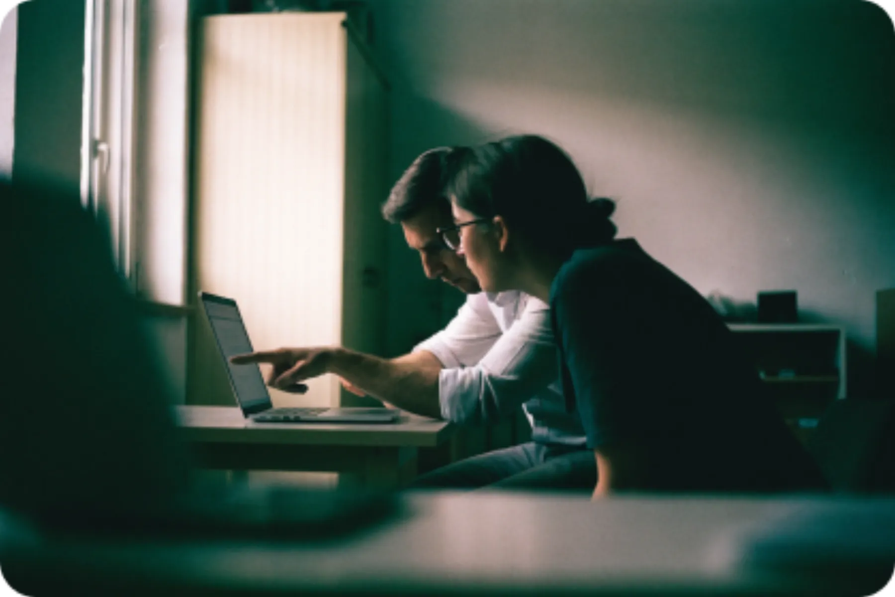 Two people at a desk in a dim room, one of them pointing at a laptop screen